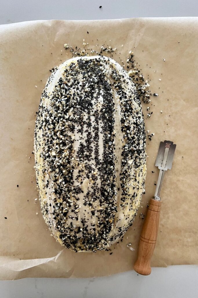 An unbaked sourdough loaf coated in Everything Bagel Seasoning. It is sitting on parchment paper with a curved blade lame. The bread has been scored to the right.