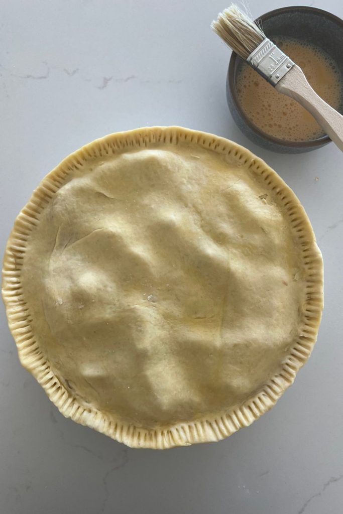 Sourdough pie crust with a crimped edge. There is a bowl of egg wash and pastry brush sitting to the right.