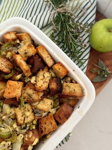 Sourdough stuffing baked in a white oven dish. Sitting on a wooden board with some herbs and an apple.