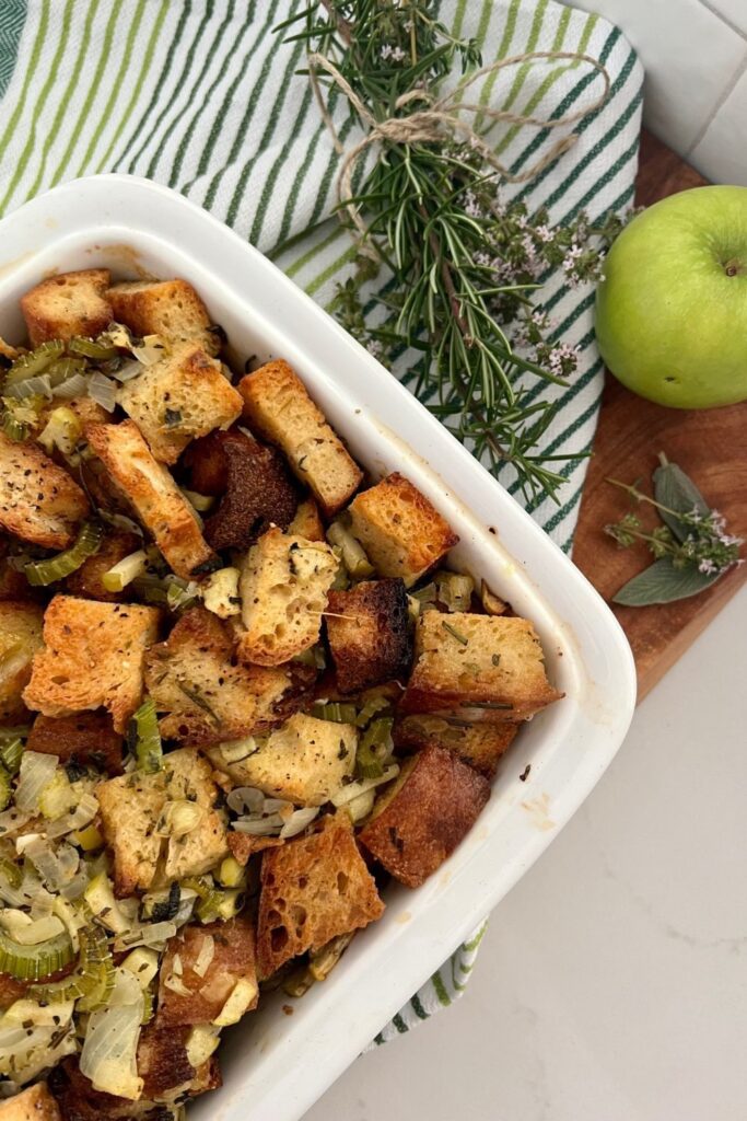 Sourdough stuffing baked in a white oven dish. Sitting on a wooden board with some herbs and an apple.