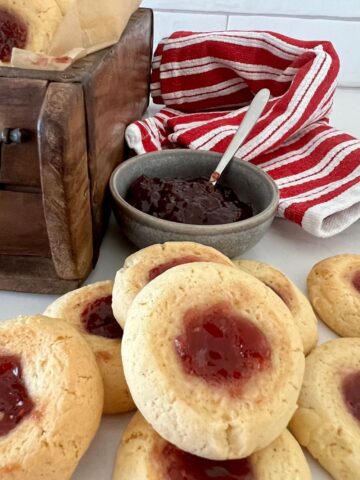 Sourdough thumbprint cookies sitting in front of a bowl of raspberry jam. There is a red and white dish cloth in the background and a wooden box.