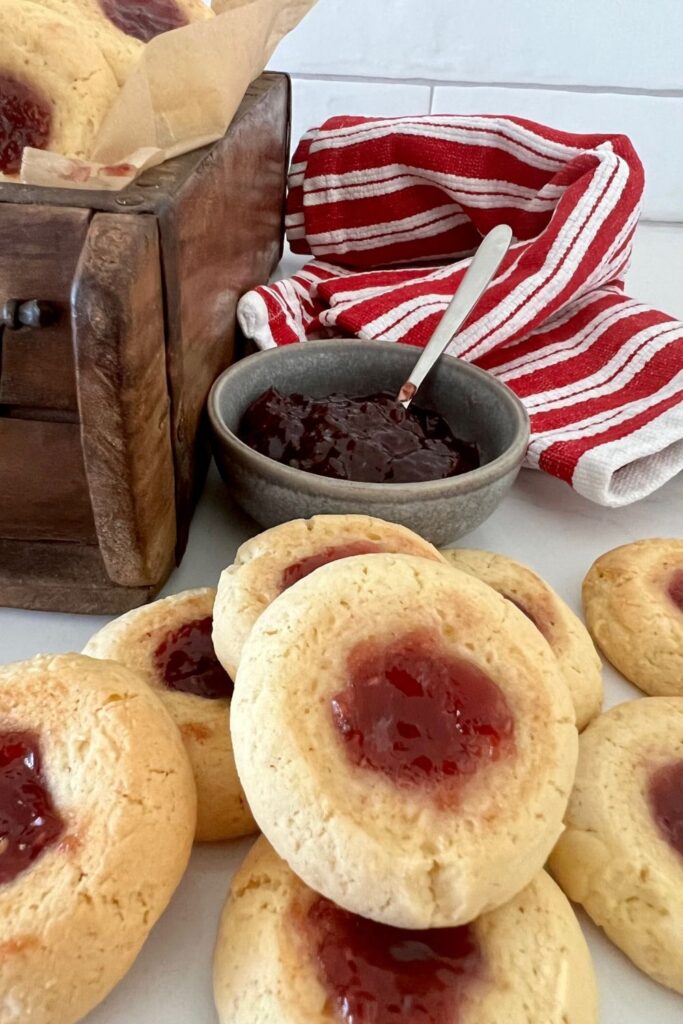 Sourdough thumbprint cookies sitting in front of a bowl of raspberry jam. There is a red and white dish cloth in the background and a wooden box.