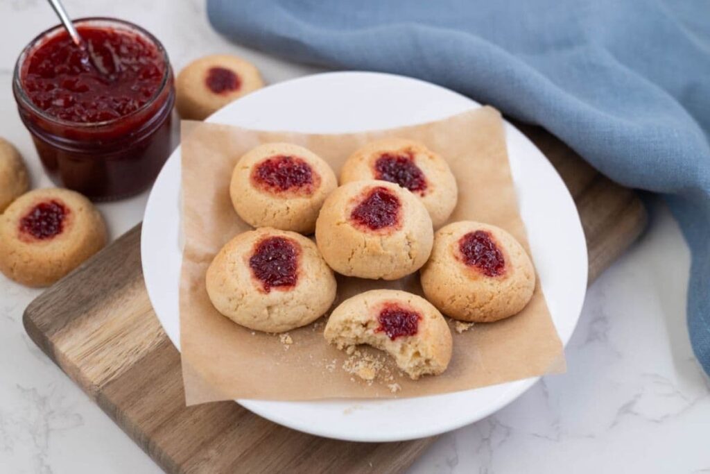6 sourdough discard thumbprint cookies sitting on a white plate. There is a jar of strawberry jam in the background.