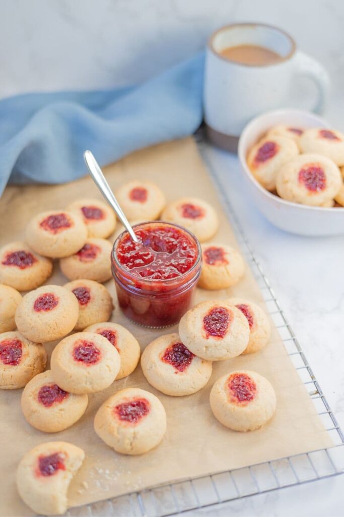 Sourdough thumbprint cookies cooling on a wire rack. There is a jar of strawberry jam in the middle of the cooling rack.