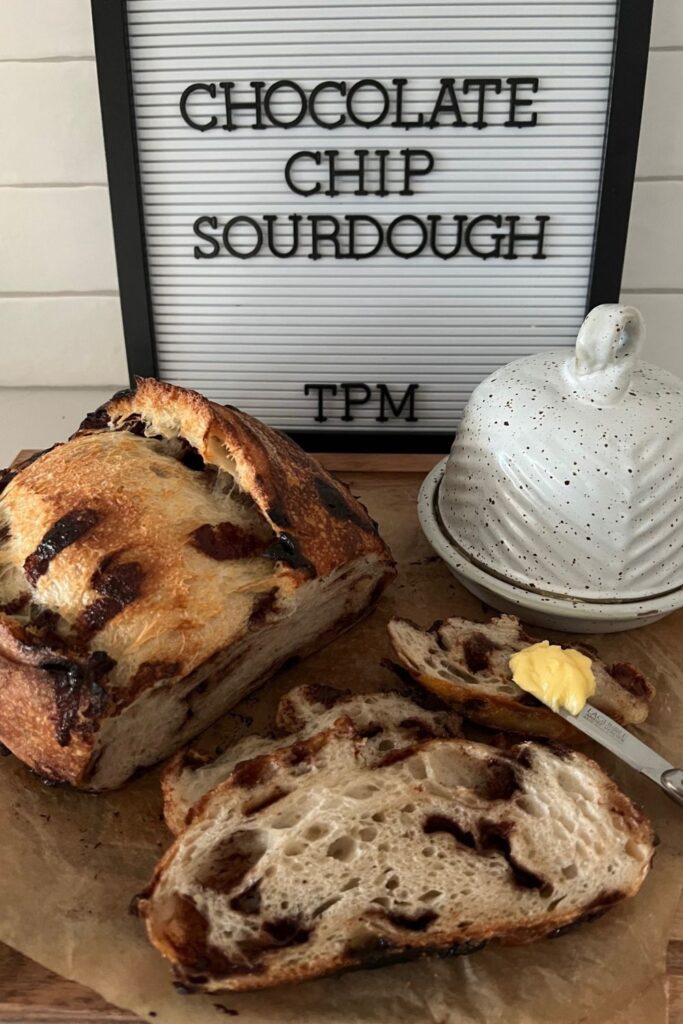 Chocolate Chip Sourdough Bread sitting in front of a sign that says "Chocolate Chip Sourdough". The loaf has been cut into pieces and there are a few slices sitting in front of the remainder of the loaf. There is a butter crock to the right and knife loaded with butter sitting one of the slices of bread.