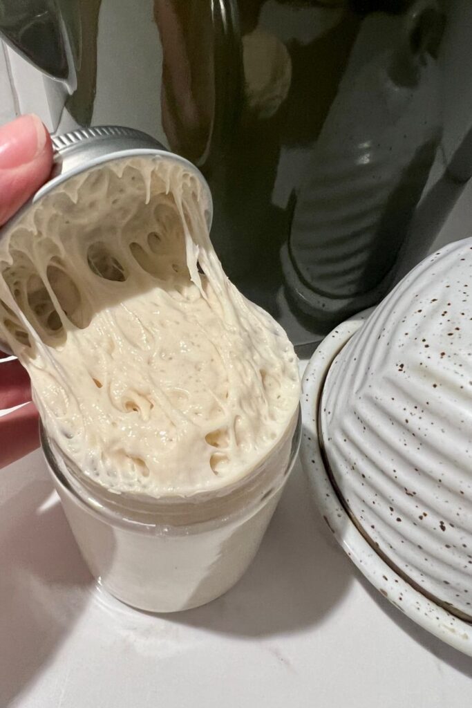 A jar of vegan sourdough starter that is overflowing the jar. The lid has been lifted to show the bubbly sourdough starter inside. There is a ceramic dish sitting to the right of the jar and a green fermentation crock behind the jar.