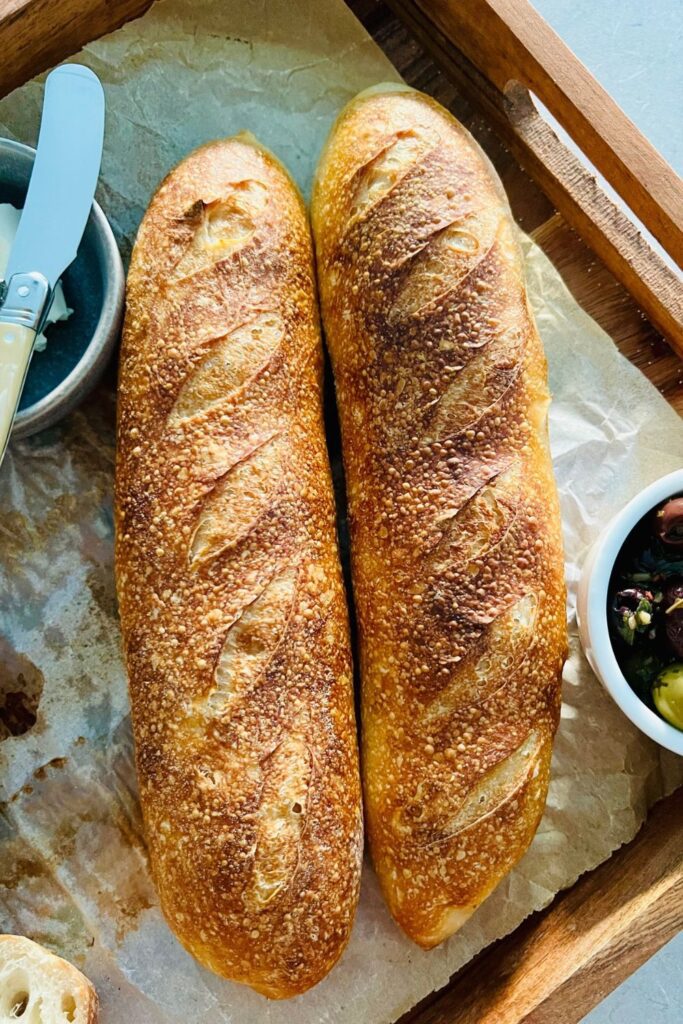 A pair of sourdough vegan baguettes sitting on a piece of parchment paper. There is a bowl of olives and a knife in the photo too.