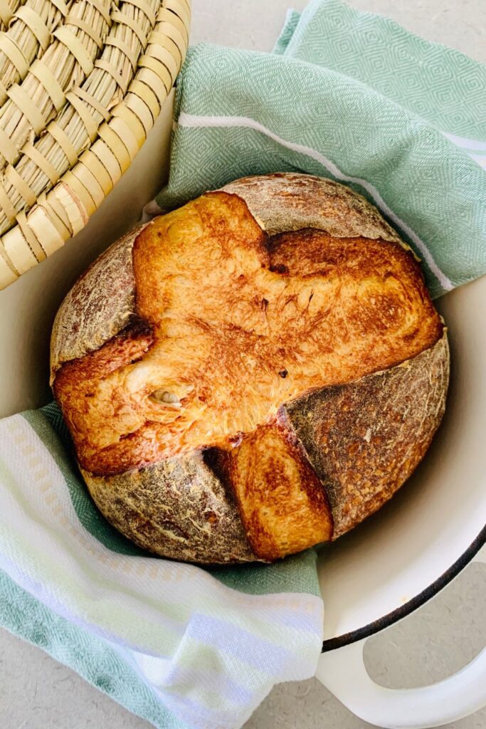 A loaf of vegan sourdough bread sitting inside a cream enamel dutch oven. There is a pale green tea towel and cane basket in the photo too.