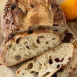 Loaf of orange cranberry sourdough bread that has been sliced. There is a whole orange sitting at the back of the bread.