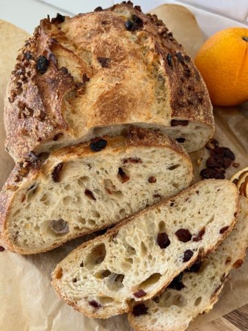 Loaf of orange cranberry sourdough bread that has been sliced. There is a whole orange sitting at the back of the bread.