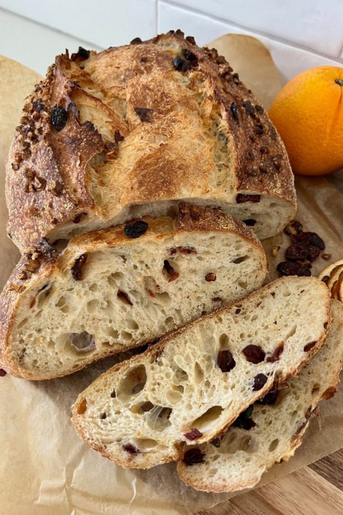 Loaf of orange cranberry sourdough bread that has been sliced. There is a whole orange sitting at the back of the bread.