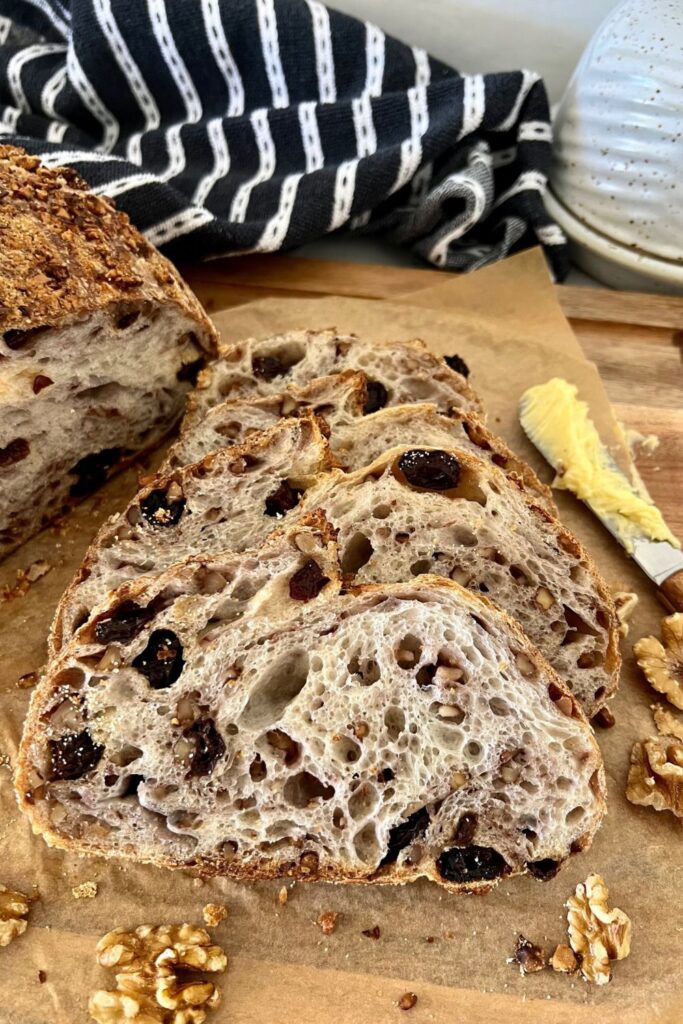 Walnut Raisin Sourdough Bread that has been sliced and arranged so you can see the "bunny" in the bread. There is a black and white dish towel sitting in the background and a wooden butter knife to the right of the sliced sourdough bread.