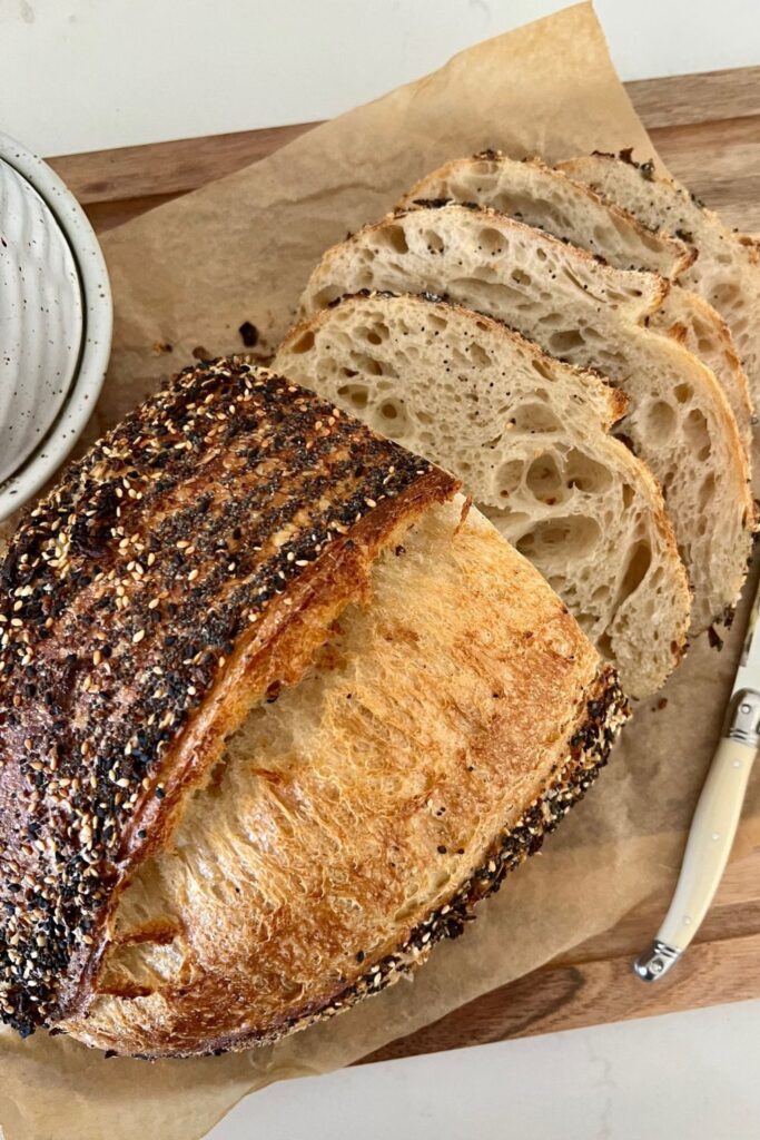 A loaf of crust sourdough bread covered in Everything Bagel Seasoning that has been sliced and arranged on a bread board.