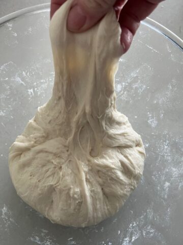 Sourdough bread dough sitting in a bowl with a hand stretching part of the dough upwards in a stretch and fold gluten development technique.