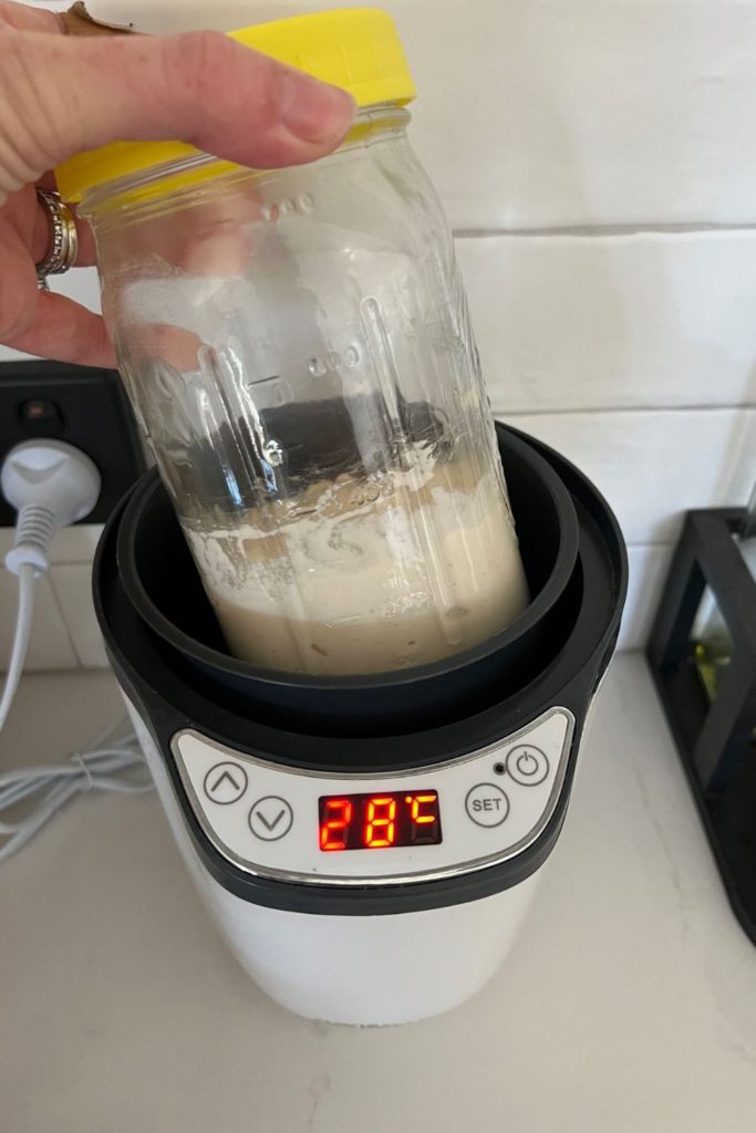 Jar of sourdough starter with yellow lid being placed into a small yogurt maker to keep it warm.