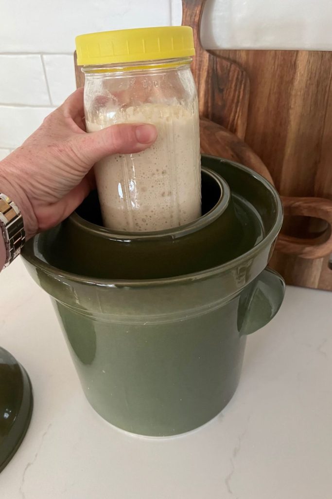 A jar of sourdough starter with a yellow lid being placed inside a fermentation crock.