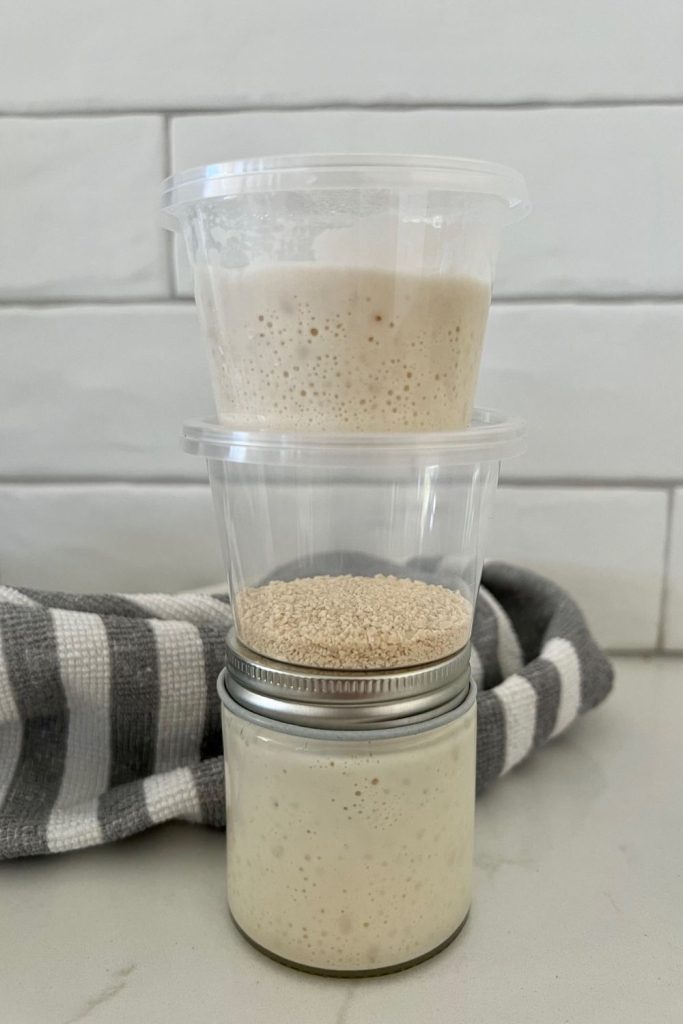A small mason jar sitting on the counter. There are two small take out containers sitting on top of the mason jar. The mason jar contains bubbly, liquid starter, the second take out container is full of dried sourdough starter and the top container is filled with bubbly sourdough starter.