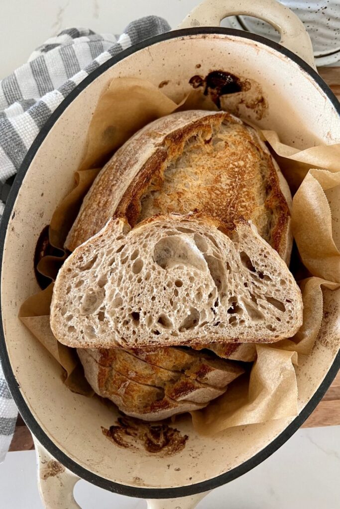 Rustic sourdough bread sitting in a cream enamel Dutch Oven. There is a slice of the bread sitting on top of the loaf to show the whole wheat crumb inside.