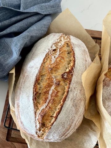 Loaf of rustic sourdough bread sitting in a piece of parchment paper on a wooden board. There is a blue dish towel on the left and you can see the edge of a second loaf of sourdough on the right.