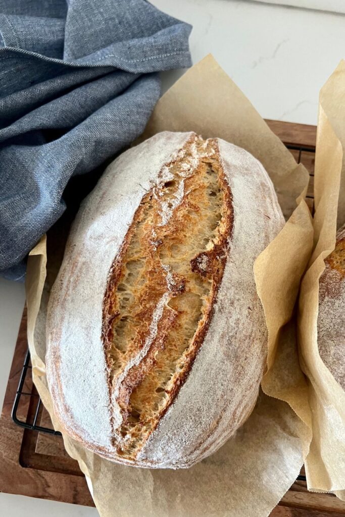 Loaf of rustic sourdough bread sitting in a piece of parchment paper on a wooden board. There is a blue dish towel on the left and you can see the edge of a second loaf of sourdough on the right.