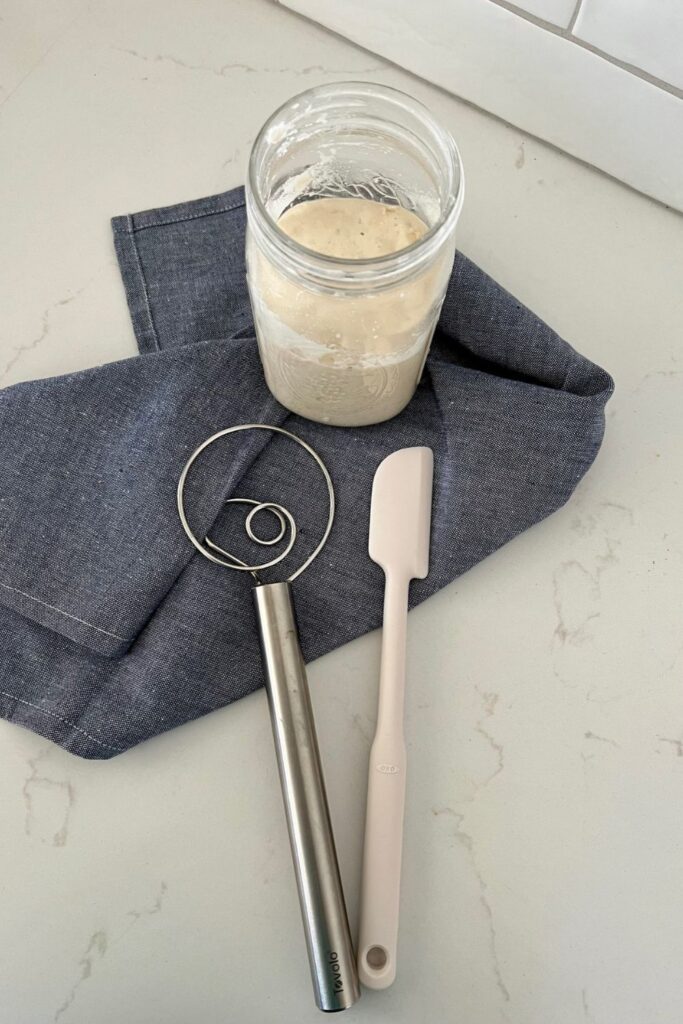 Jar of sourdough starter sitting on a denim blue dish cloth. There is a silver Danish Whisk and white jar spatula sitting in front of the jar.