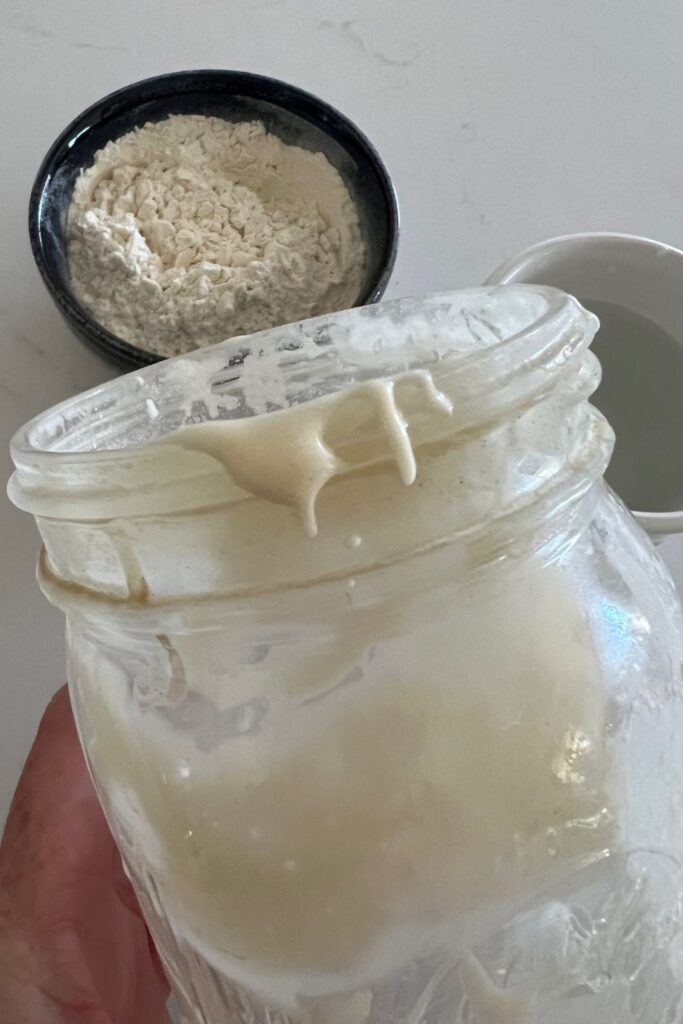 Close up of a sourdough starter jar with excess sourdough starter sitting around the rim of the jar. There is a bowl of flour in the background.