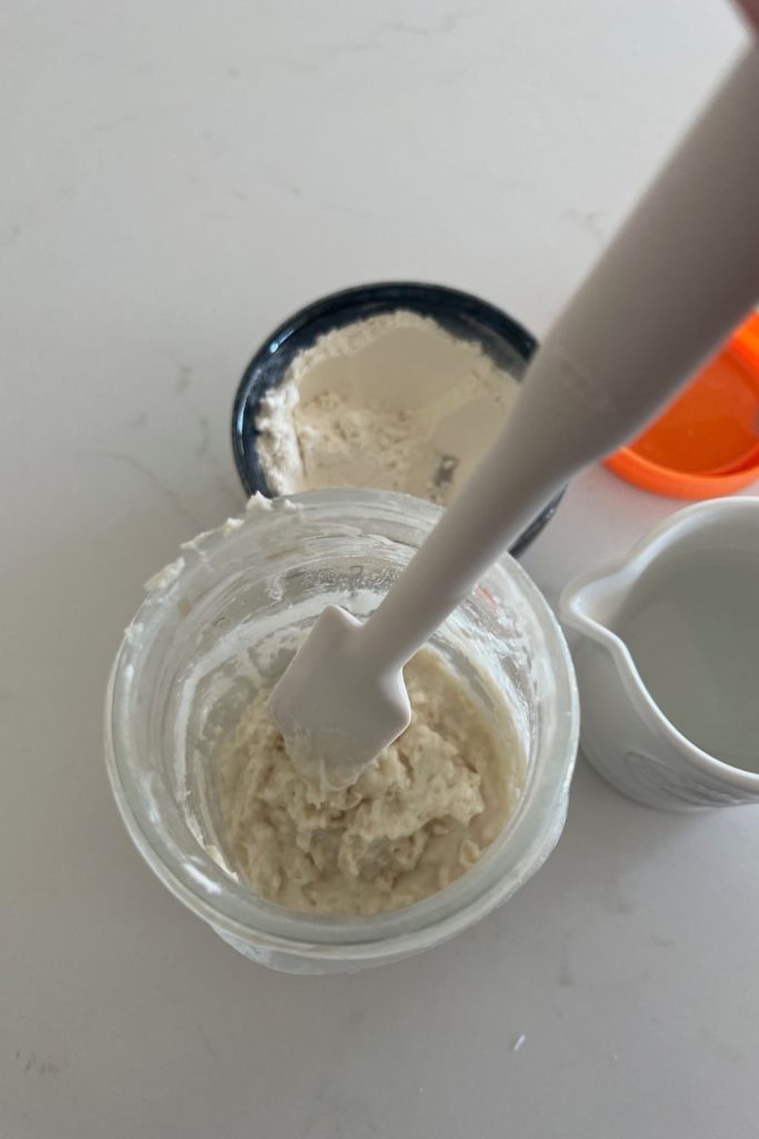White jar spatula being used to scrape down the sides of the sourdough starter jar.