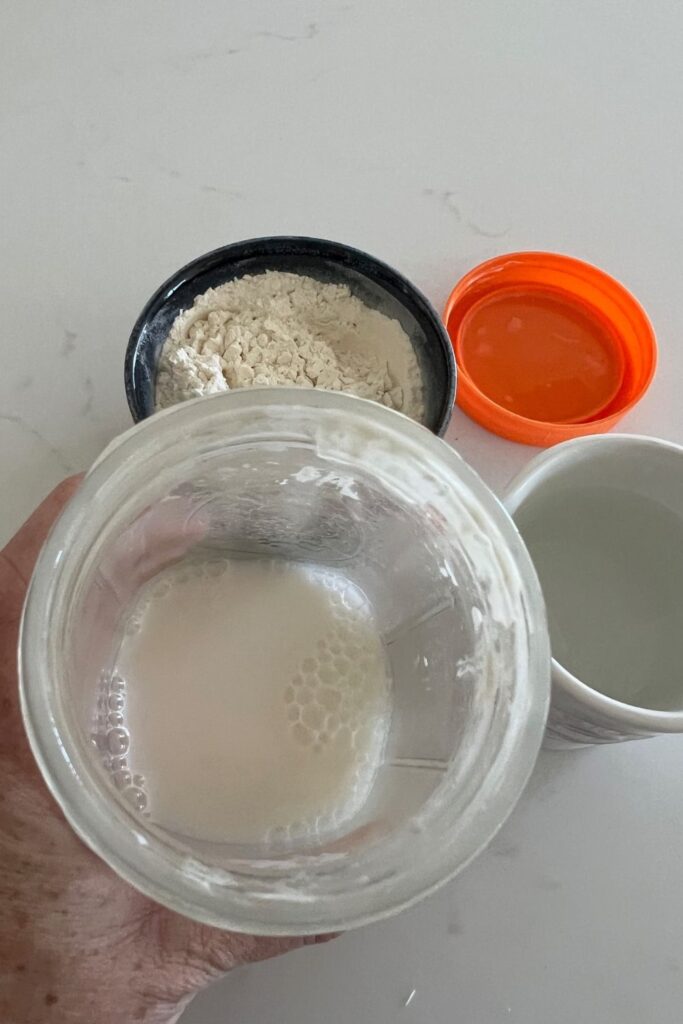 Wide mouth mason jar that has a watery sourdough starter in it. There is a bowl of flour, orange jar lid and jug of water in the background.