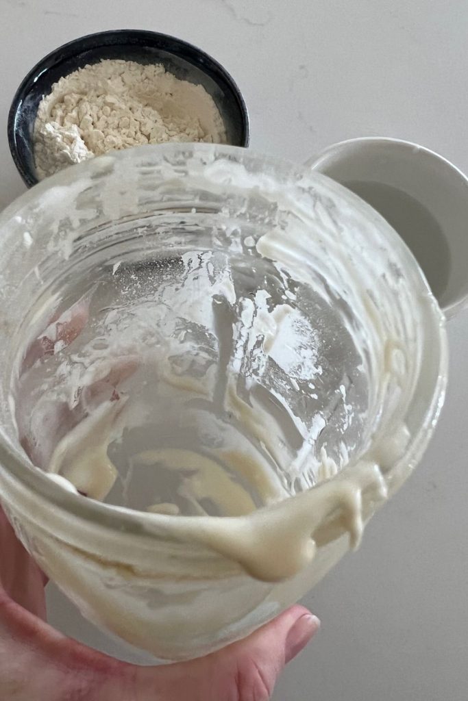 Jar of sourdough starter that has been emptied out but is still dirty. There is some sourdough starter around the edges of the jar and there is a bowl of flour and jug of water in the background of the photo.