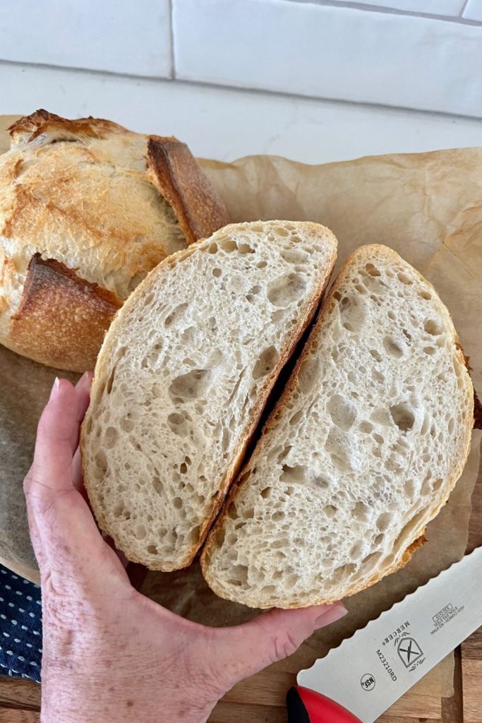 Small loaf of sourdough that has been sliced in half and is being held together to show the crumb. There is a red Mercer knife in the bottom right hand corner and another small loaf in the background.