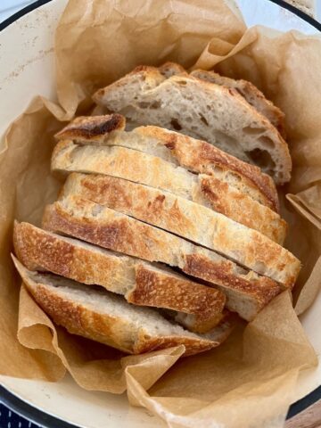 Small loaf of sourdough bread that has been sliced sitting in a cream enamel Dutch Oven surrounded by parchment paper.