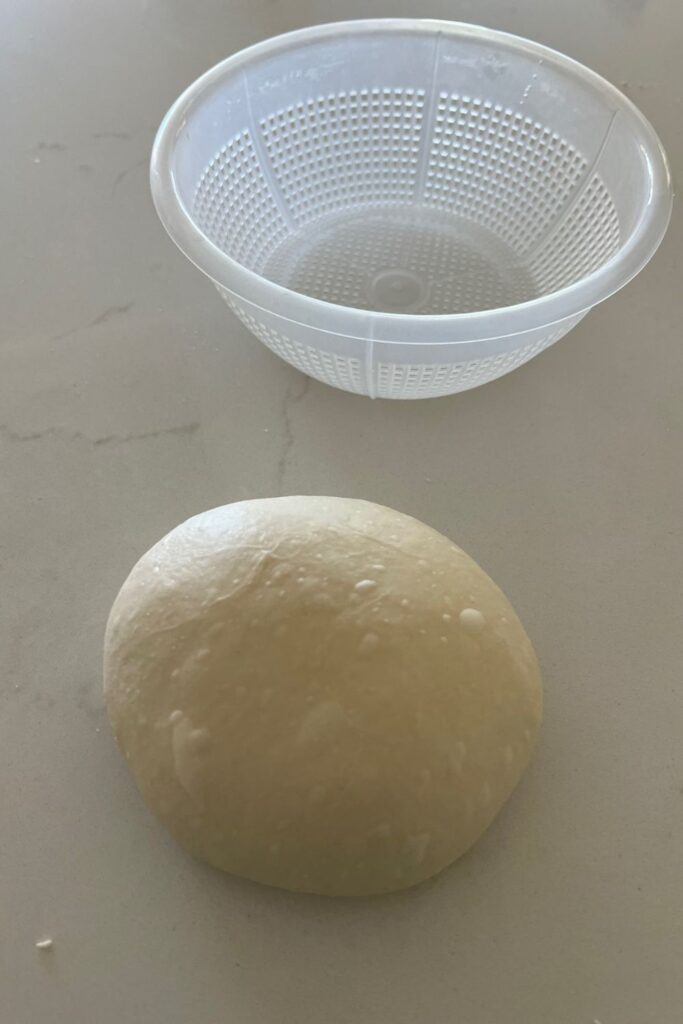 Small batch sourdough that has been shaped into a boule. There is a ricotta basket above it.