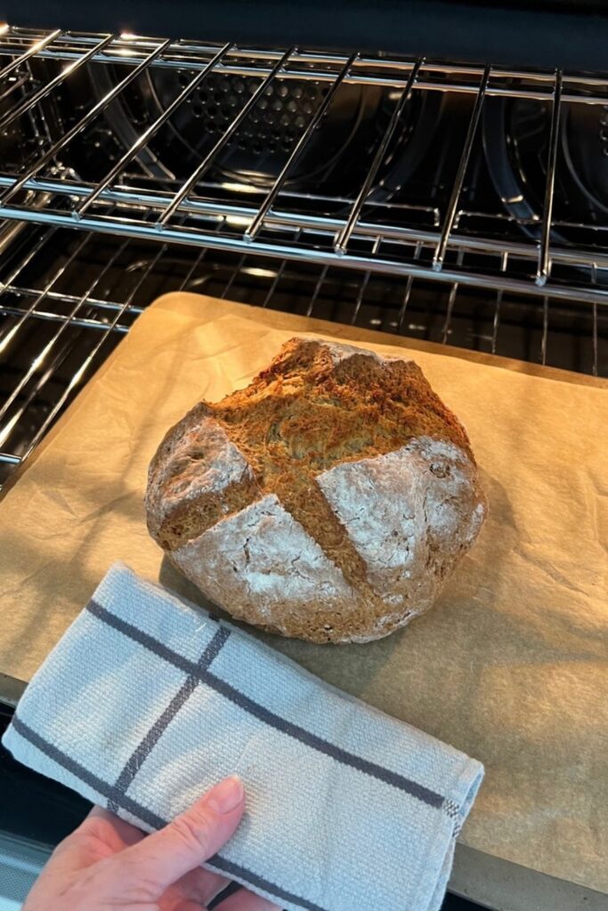 A loaf of Irish Sourdough Soda Bread being pulled out of the oven. The bread is golden brown and there is a grey dish towel holding the hot oven tray.