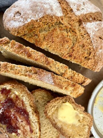 Loaf of sourdough Irish soda bread sitting on a board. There are a few slices of bread with butter and jam and a blue dish towel in the background.