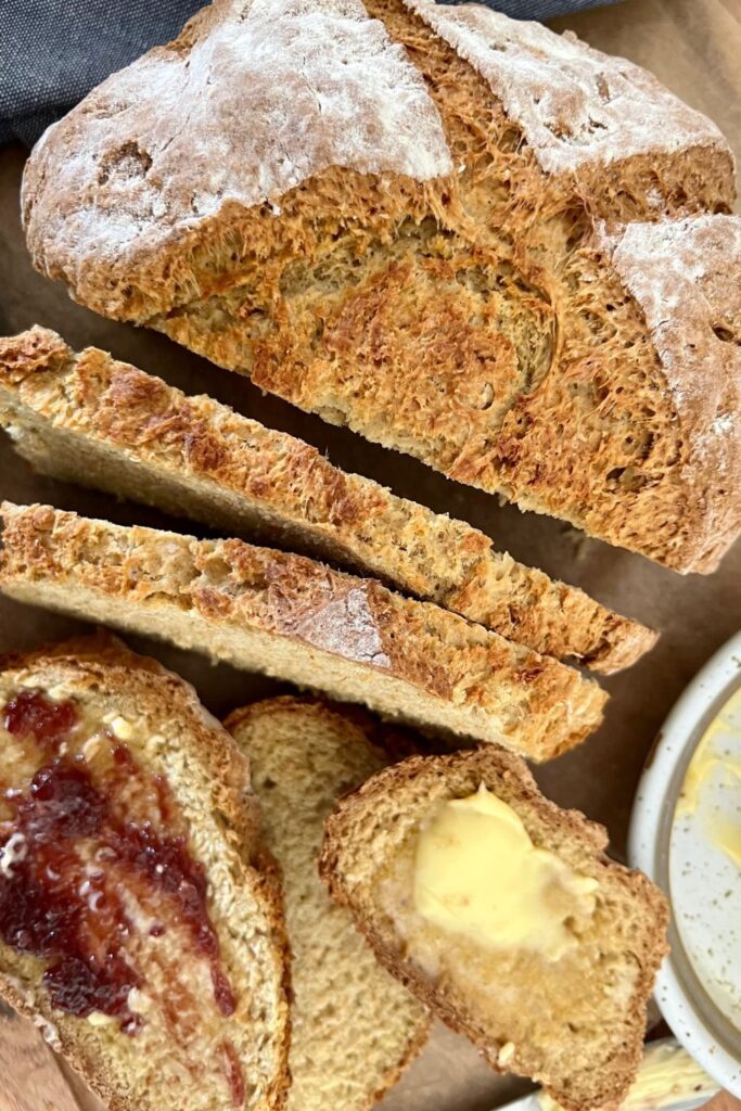 Loaf of sourdough Irish soda bread sitting on a board. There are a few slices of bread with butter and jam and a blue dish towel in the background.