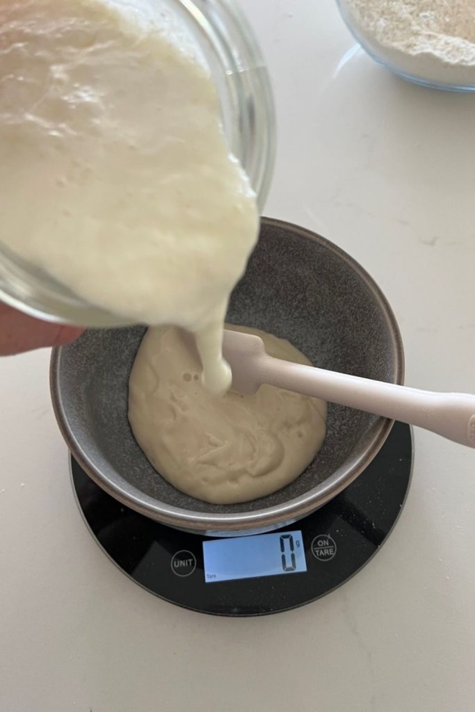 Pouring buttermilk into a bowl of sourdough starter. The bowl is sitting on a set of black digital scales and there is a white spatula in the bowl.