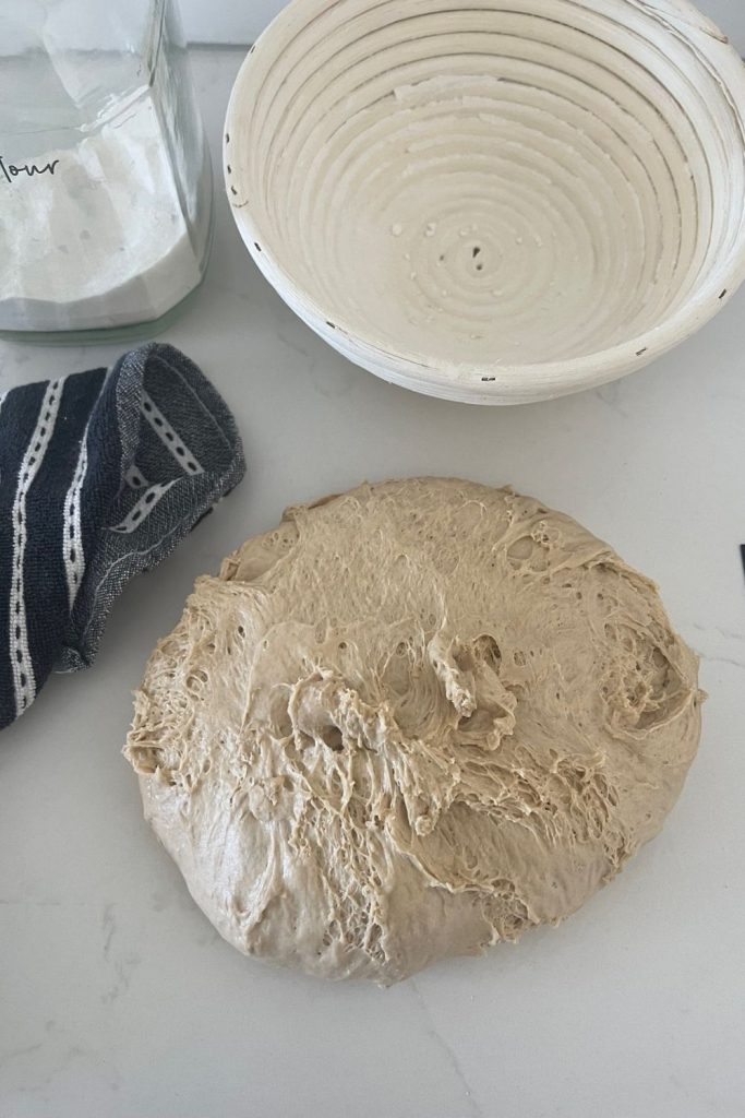 Sourdough rye bread dough tipped out of the bowl and onto the counter. There is a black dish towel, banneton and jar of rice flour in the photo too.