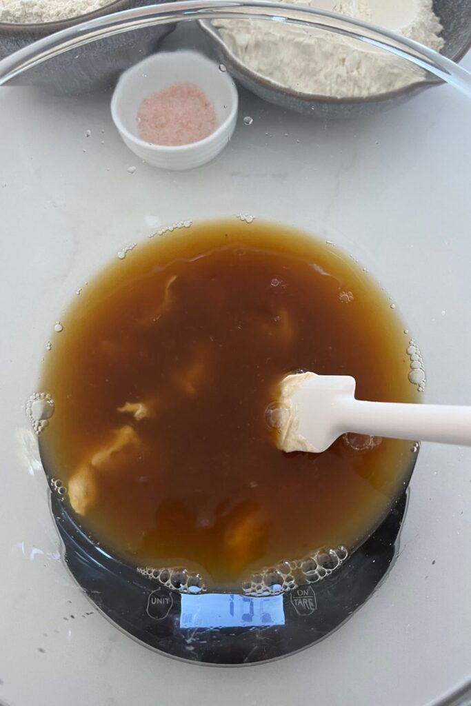 A glass bowl with sourdough starter, water and molasses added into it and stirred together with a white rubber spatula. There is a black scale under the bowl and a small white bowl of pink salt in the background.