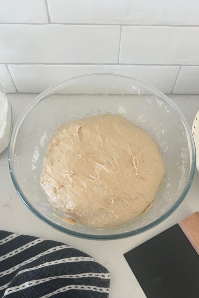 Sourdough rye dough sitting in a glass bowl with a black and white dish towel and black dough scraper next to it. The dough is puffy and ready to be shaped.