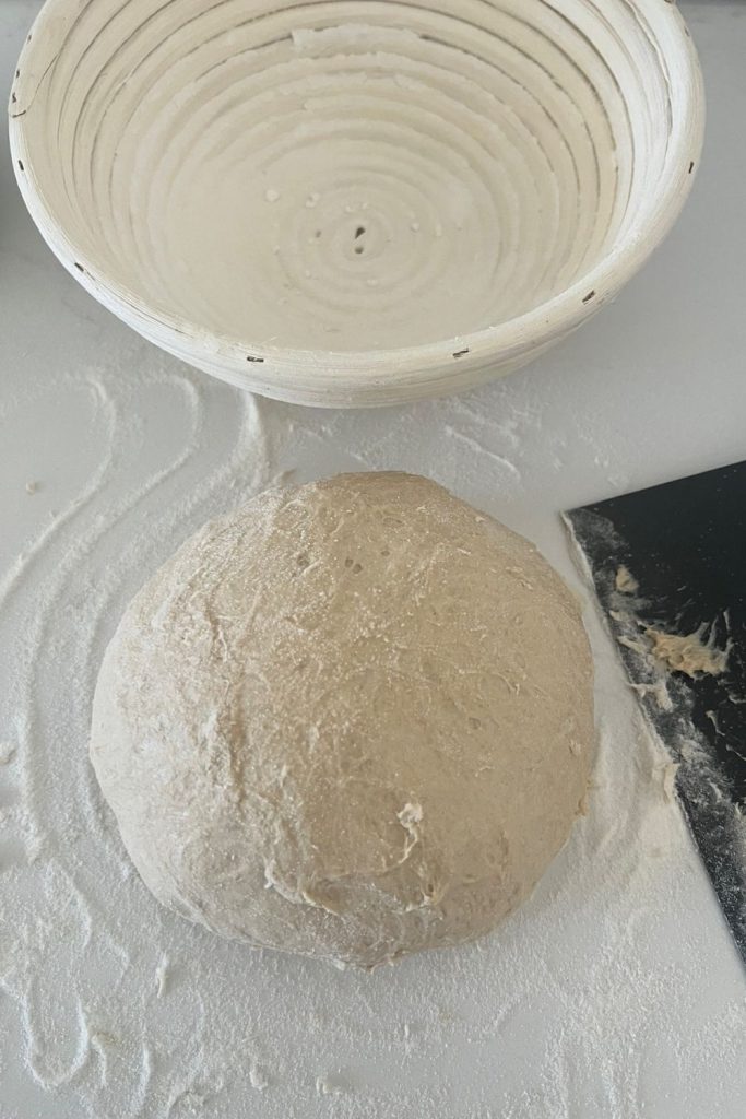Sourdough rye that has been shaped into a boule. There is a round banneton sitting above the dough and lots of rice flour around it. There is sticky dough stuck to the black dough scraper.