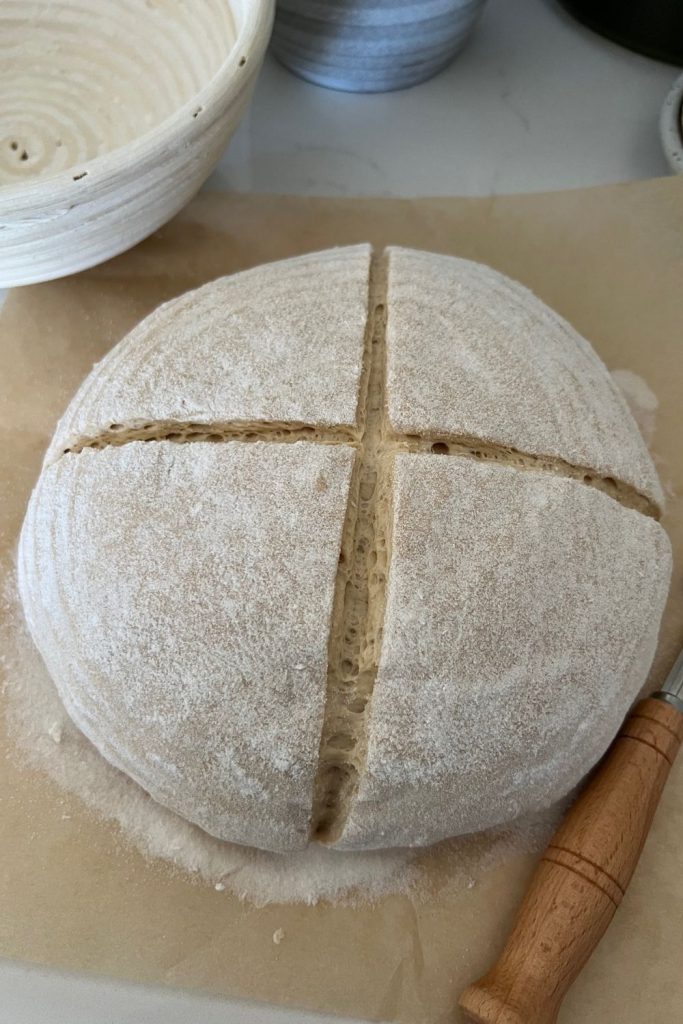 Sourdough rye boule that has been tipped out onto a piece of parchment paper. The dough has been scored with a cross. There is a wooden handled lame in the bottom left hand corner of the picture.