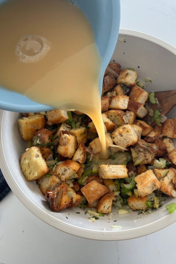 Cubed leftover sourdough bread tossed with herbs and a blue jug pouring egg mixture over the bread cubes.