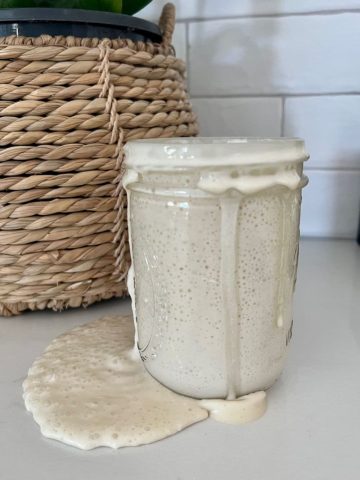 An overfed sourdough starter that has overflowed the jar that it is in. The sourdough starter is dribbling down the side of the jar and onto the counter making a big mess. There is a cane basket with a plant in the background behind the jar of sourdough starter.