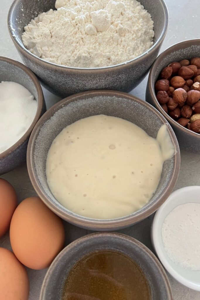 A bowl of sourdough discard surrounded by bowls of flour, nuts, eggs.