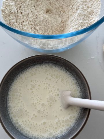 How to convert yeast recipe to sourdough - small blue bowl with bubbly sourdough starter and a white jar spatula. There is a glass bowl of flour in the background.