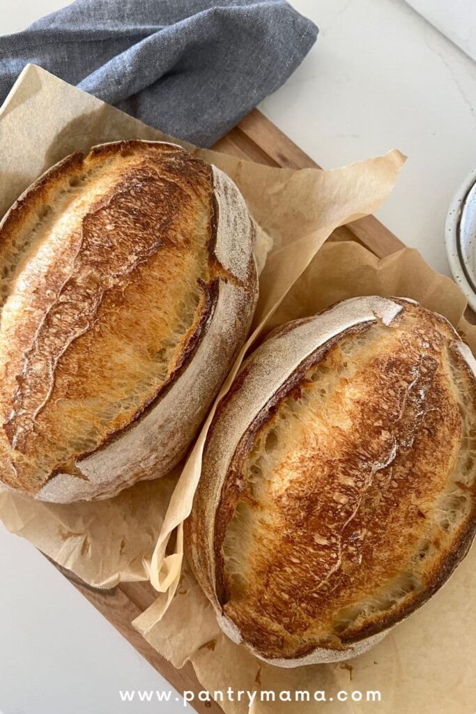 2 loaves of sourdough bread sitting side by side on a wooden board. There is a blue dish towel to the left of the loaves.