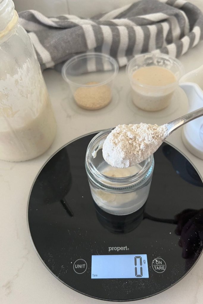 A small mason jar sitting on a black kitchen scale. The jar is filled with water and sourdough starter and there is a spoon of rye flour about to be placed into the jar. You can see a jug of water and big jar of sourdough starter in the background.