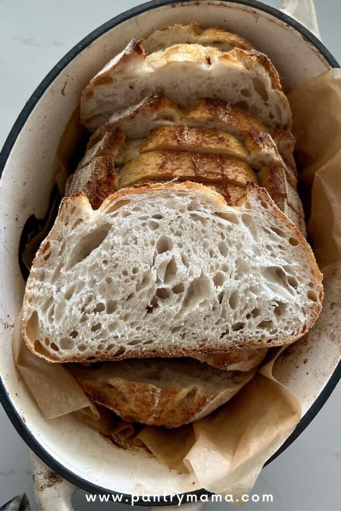 Loaf of overnight sourdough bread sliced and placed back into the Dutch oven. There is a slice laid on top of the loaf so you can see the crumb.