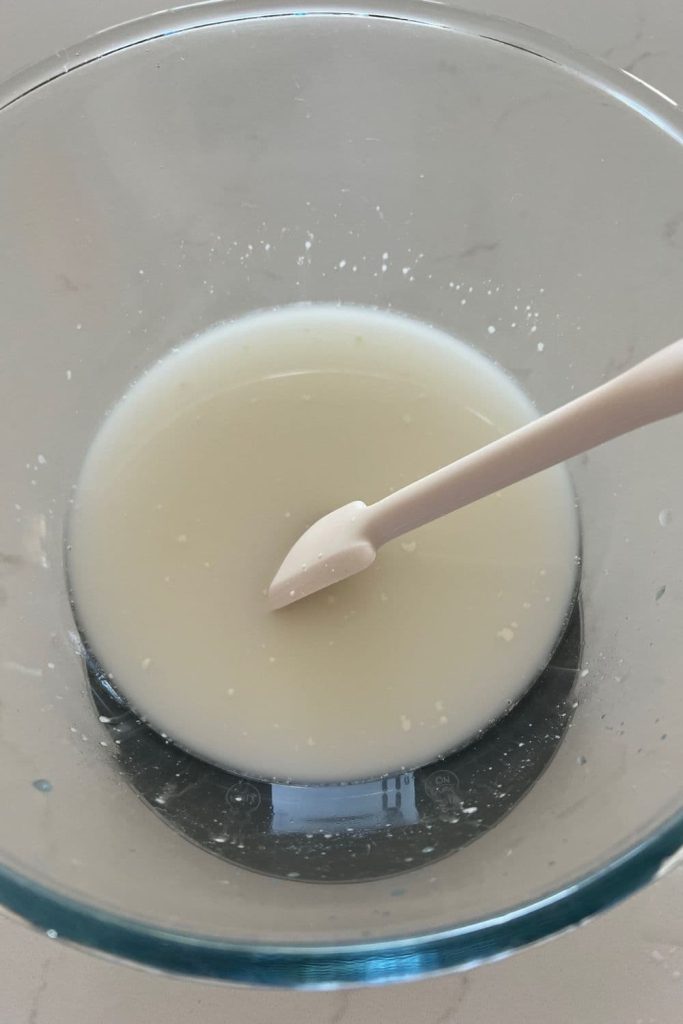 Water and sourdough starter mixed together using a white jar spatula. The glass bowl is sitting on a set of kitchen scales.