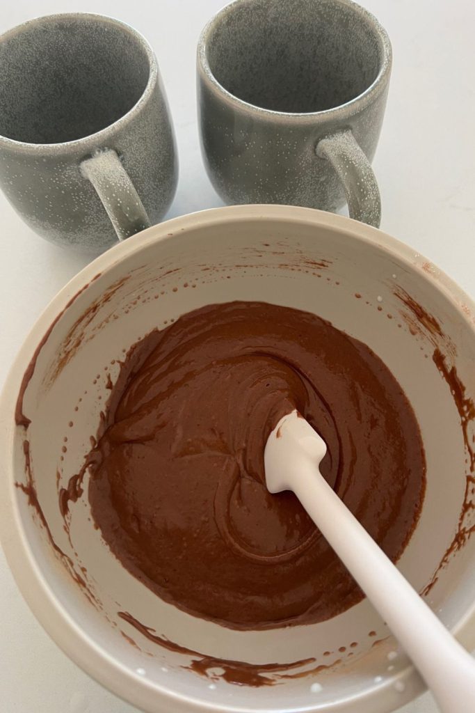 Sourdough Chocolate cake batter mixed in a bowl with a white spatula. There are two stone ware mugs sitting at the back of the bowl.